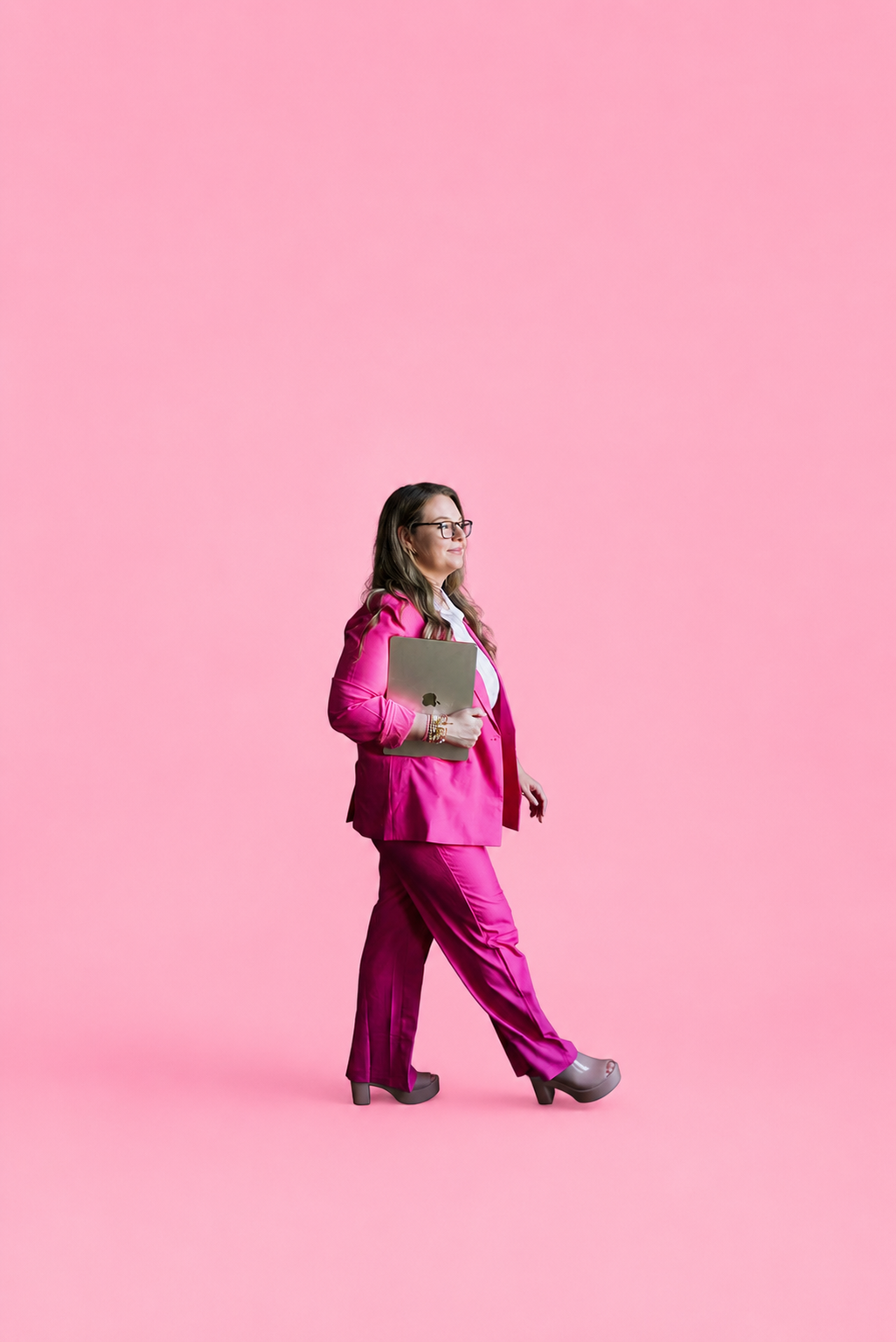 Katie Coleman walking confidently against a pink backdrop holding a laptop